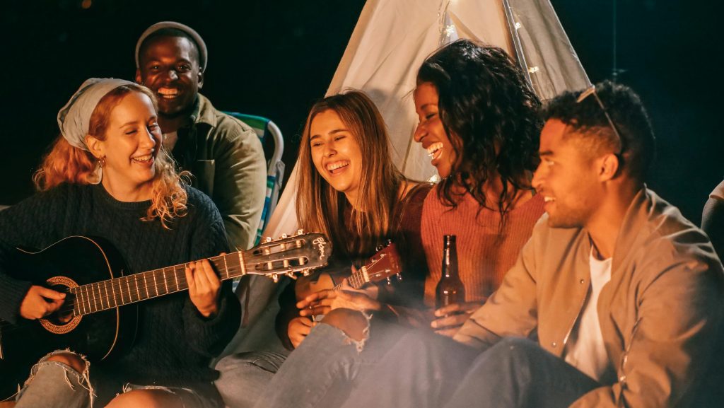 A diverse group of friends playing guitar and ukulele by a campfire at night, having fun and socializing.